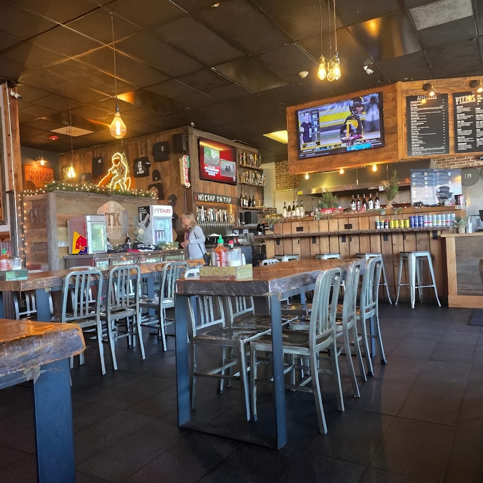 A rustic restaurant interior with wooden tables, metal chairs, a bar, and TVs showing sports.