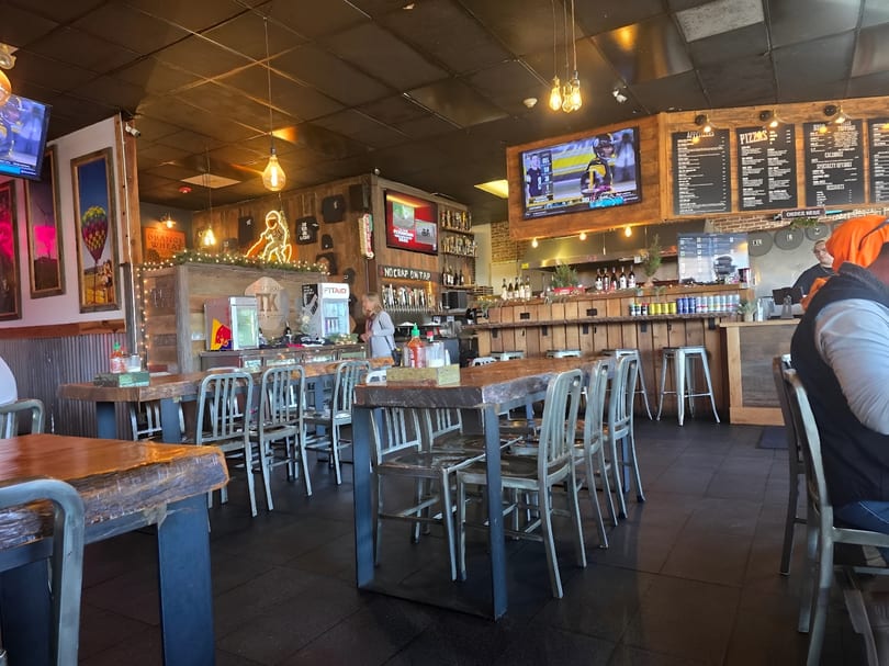A rustic restaurant interior with wooden tables, metal chairs, a bar, and TVs showing sports.