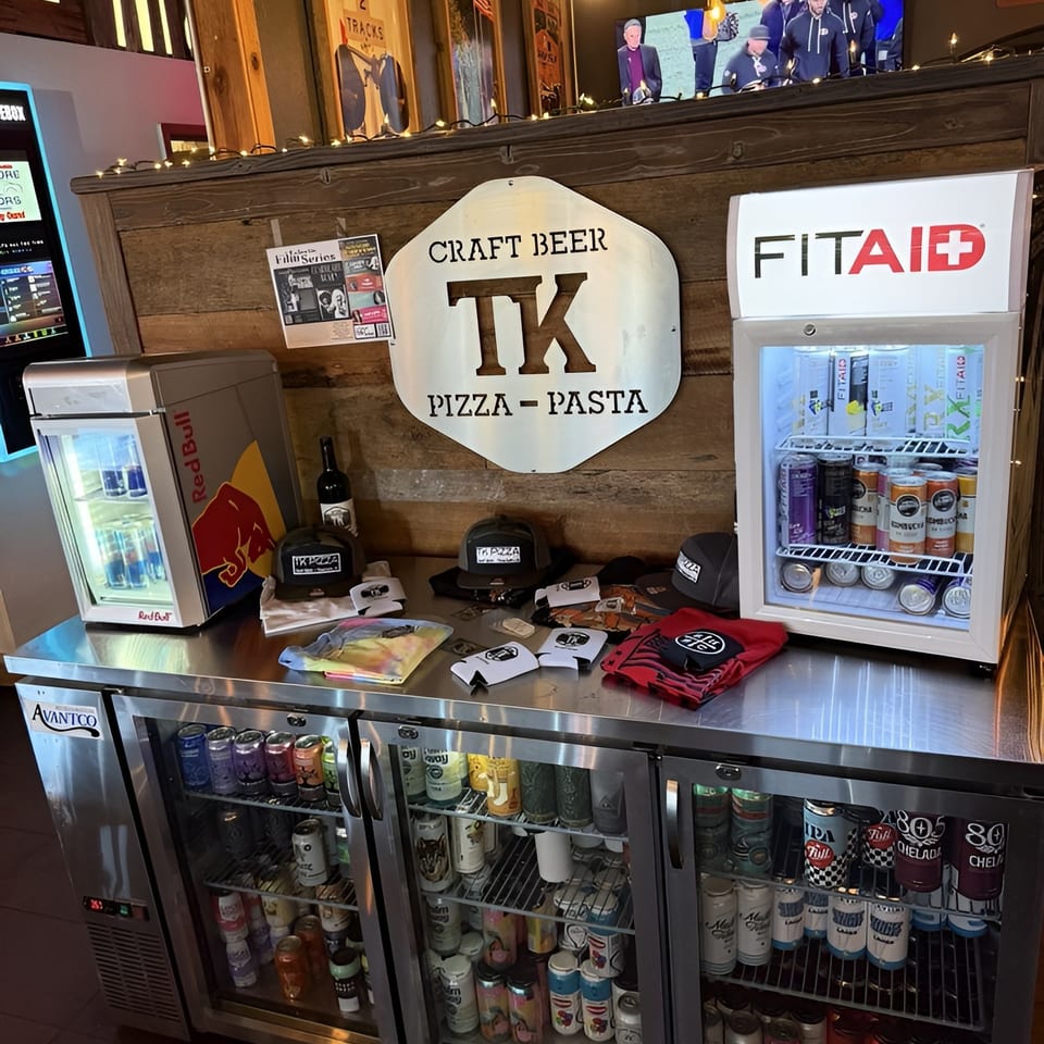 Restaurant counter with TK Pizza sign, merchandise, and refrigerators stocked with beverages.
