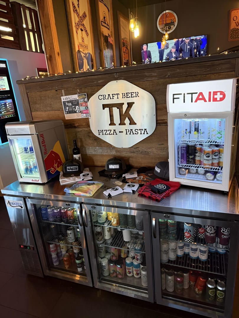 Restaurant counter with TK Pizza sign, merchandise, and refrigerators stocked with beverages.