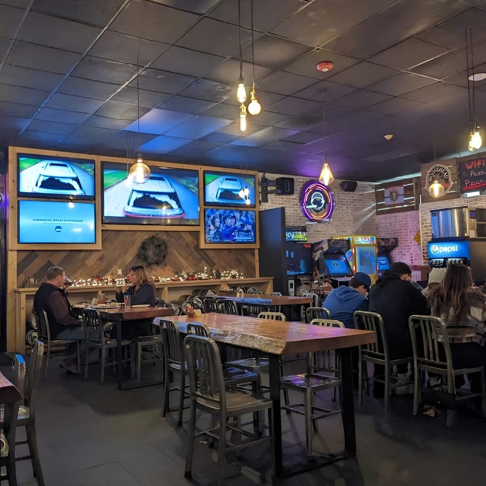 Diners seated at tables in a dimly lit restaurant with multiple TVs showing racing and arcade games.