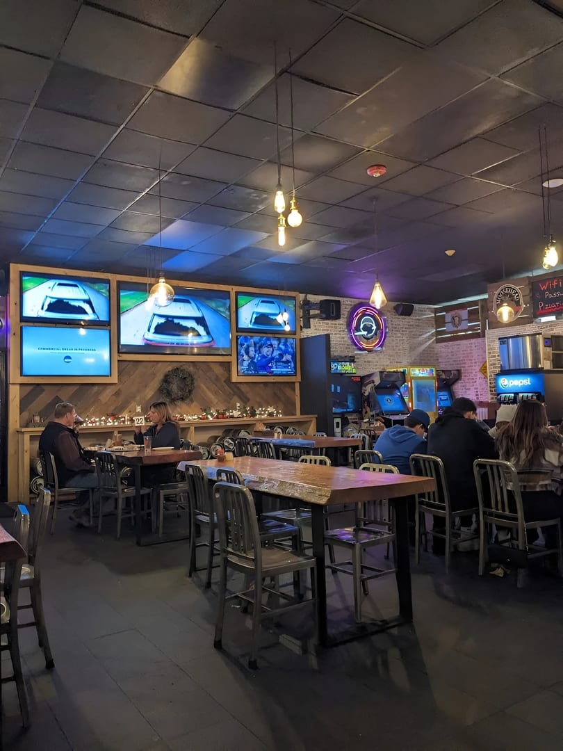 Diners seated at tables in a dimly lit restaurant with multiple TVs showing racing and arcade games.