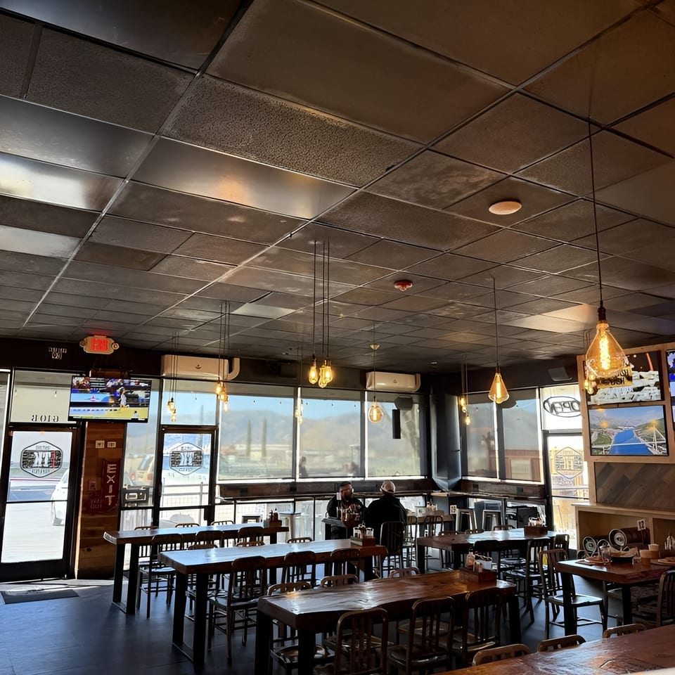 Interior of a restaurant with dark tables, chairs, and a few patrons, bathed in natural light from large windows.