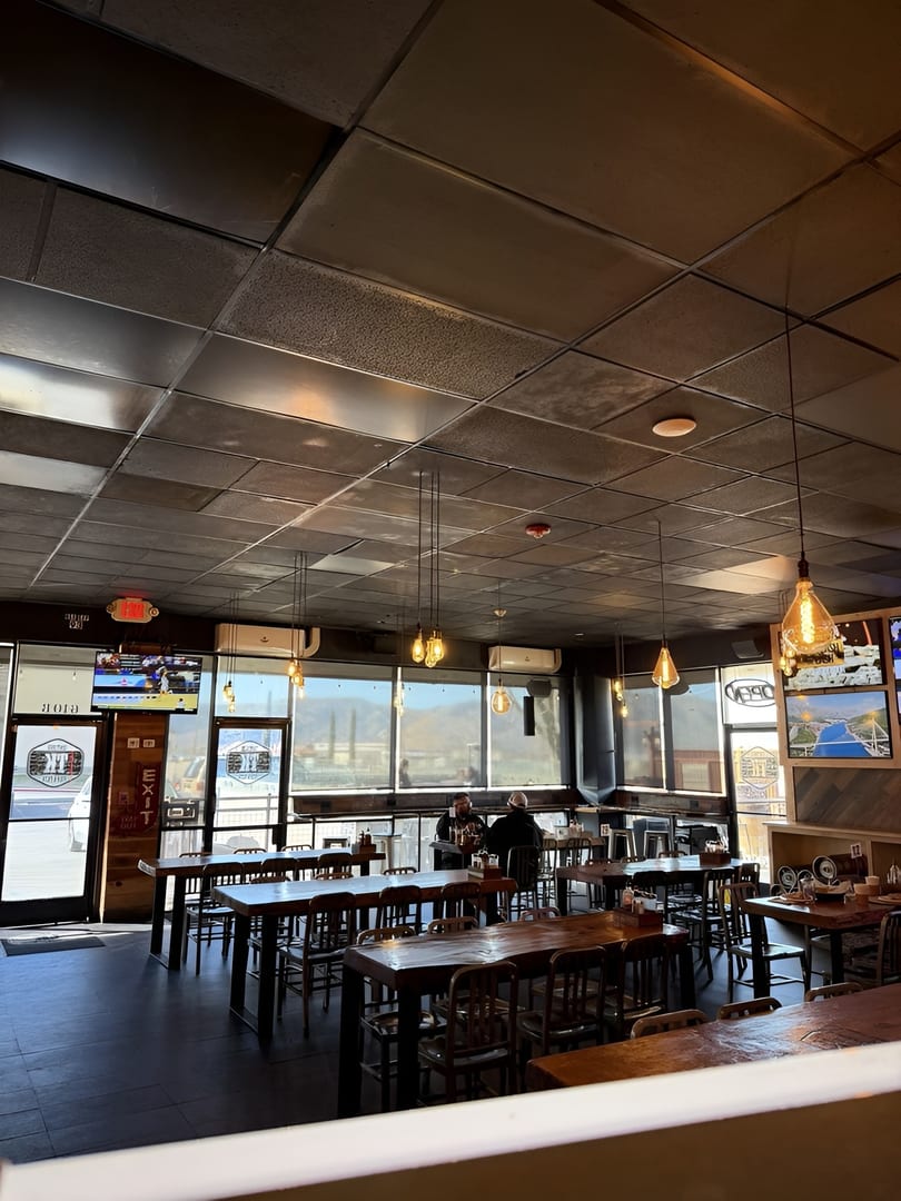 Interior of a restaurant with dark tables, chairs, and a few patrons, bathed in natural light from large windows.