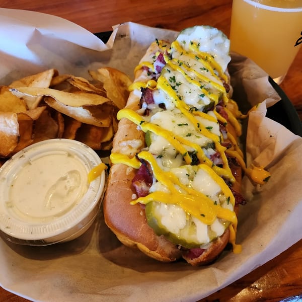 Loaded hot dog with melted cheese, pickles, mustard, bacon, and parsley, served with chips and dip.