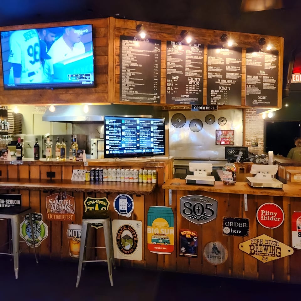 A rustic bar area with wooden counters, menu boards, beer signs, and a TV showing a baseball game.