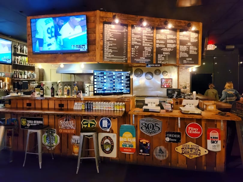A rustic bar area with wooden counters, menu boards, beer signs, and a TV showing a baseball game.
