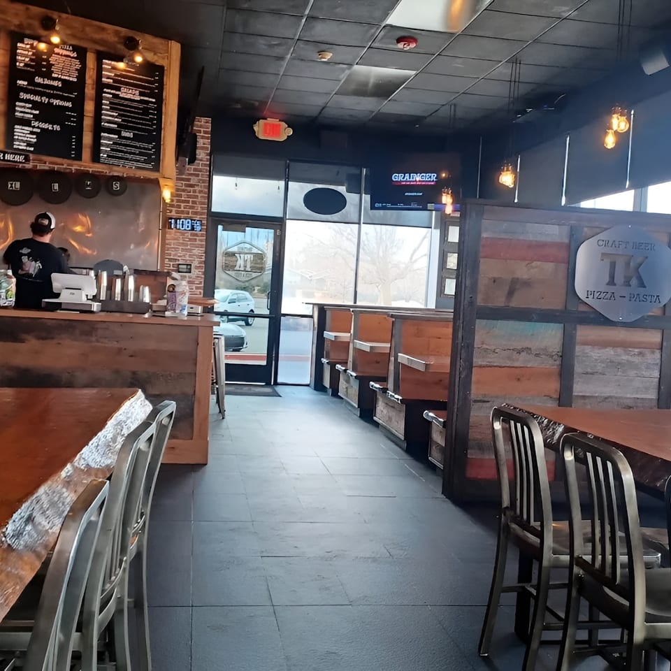 Interior of a TK Pizza and Pasta restaurant with wooden tables, booths, and a counter with a worker.